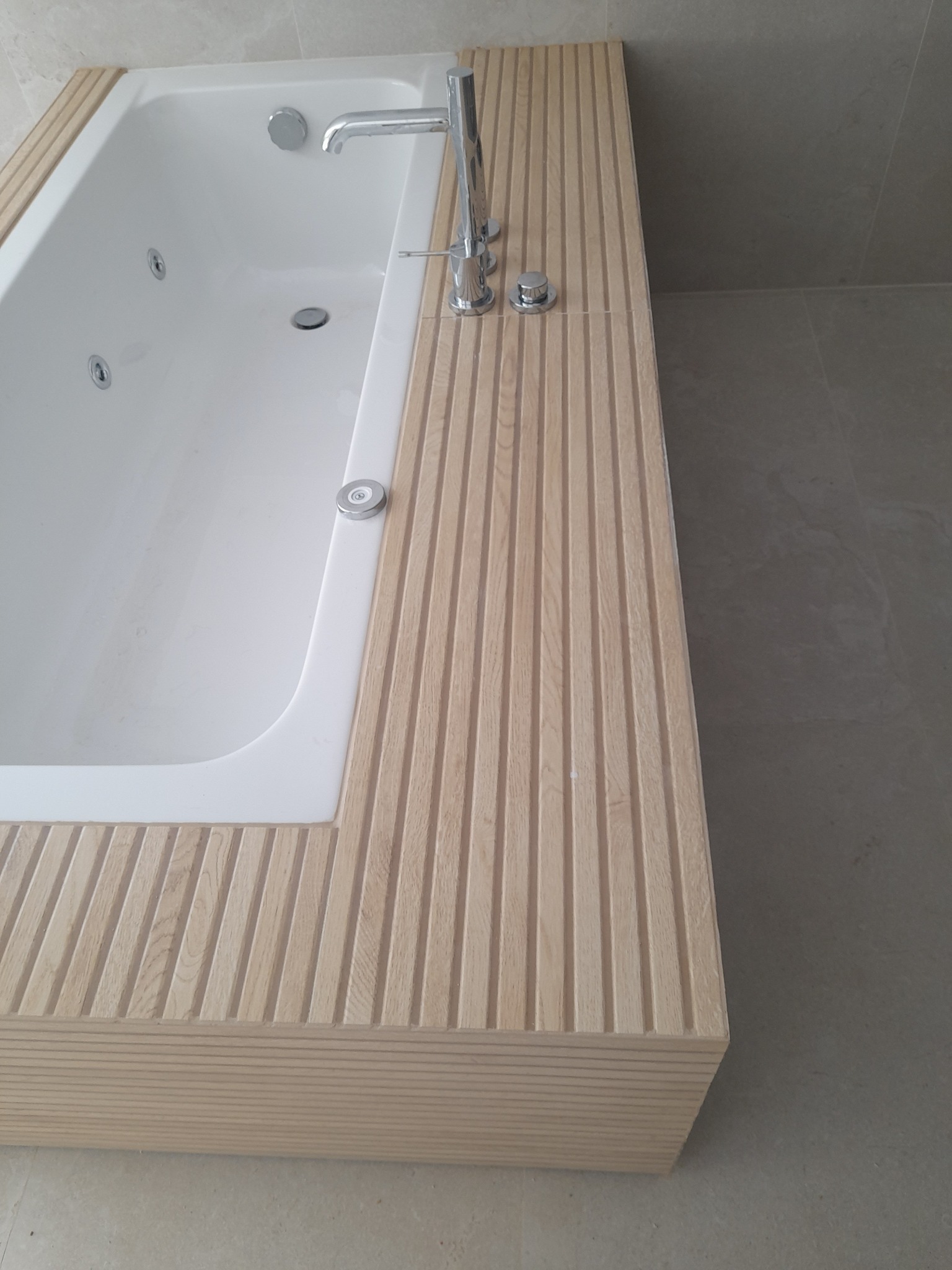 Nowoczesna wanna z hydromasażem obudowana jasnym drewnem z frezowanymi pionowymi rowkami, bateria umywalkowa chromowana, szara płytka podłogowa i ścienna w tle.