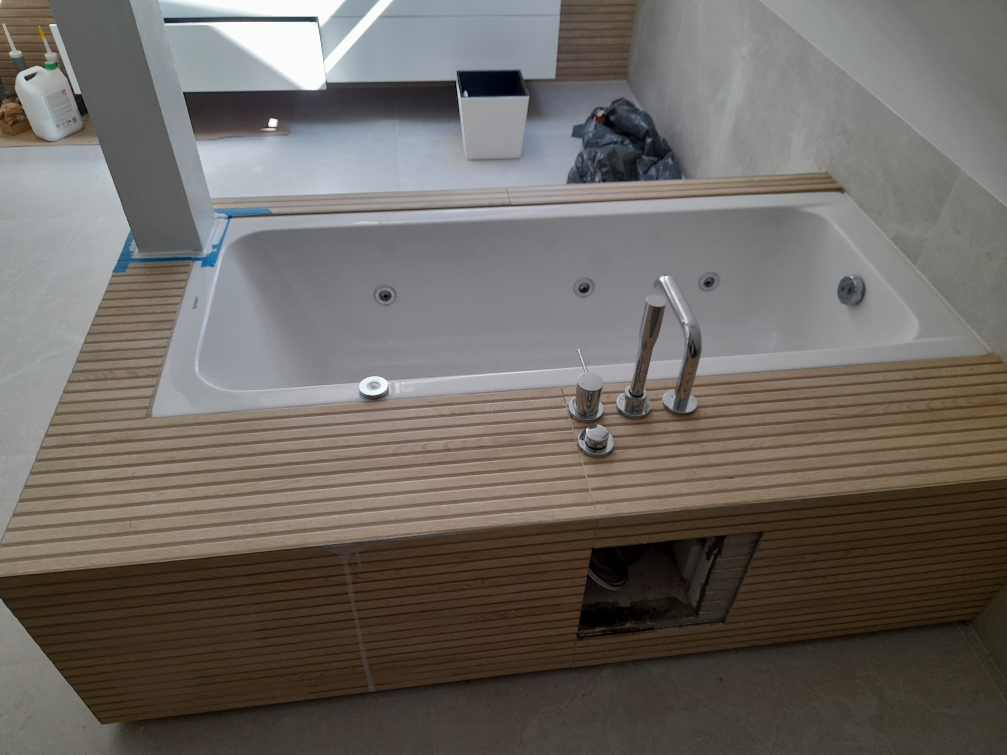 Biała wanna z hydromasażem, obudowana drewnianymi panelami, z widoczną armaturą i otworem rewizyjnym, w trakcie prac wykończeniowych w łazience.