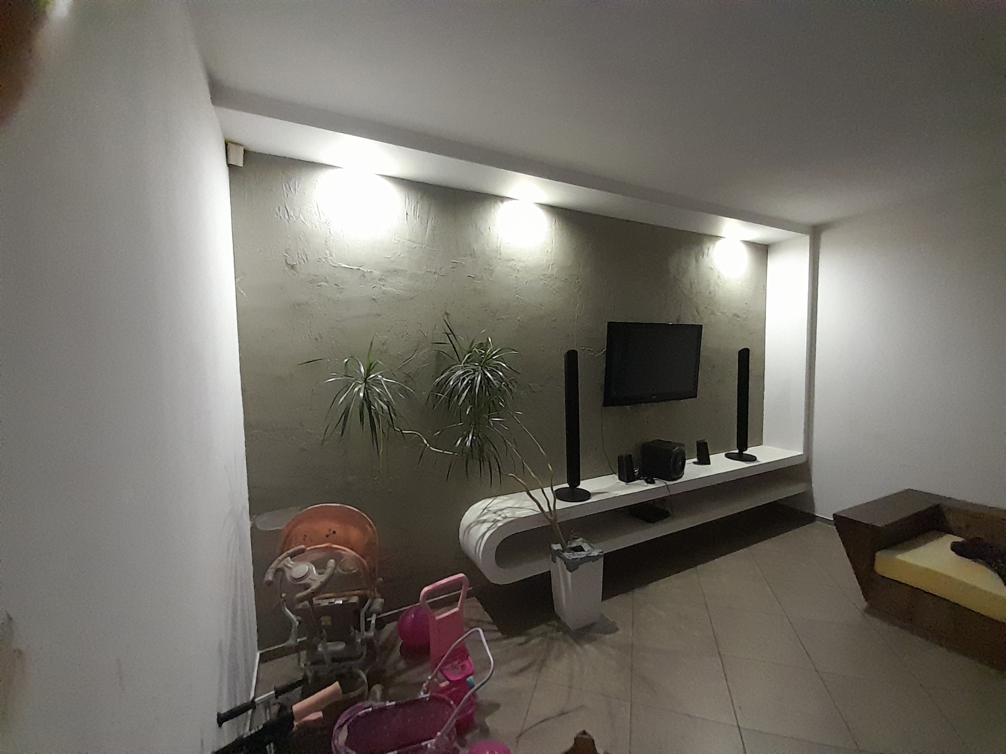 Ściana w salonie z dekoracyjną strukturą, oświetlona listwą LED, z roślinami i meblami, w tle sprzęt audio-video. Na pierwszym planie dziecięce wózki i zabawki.