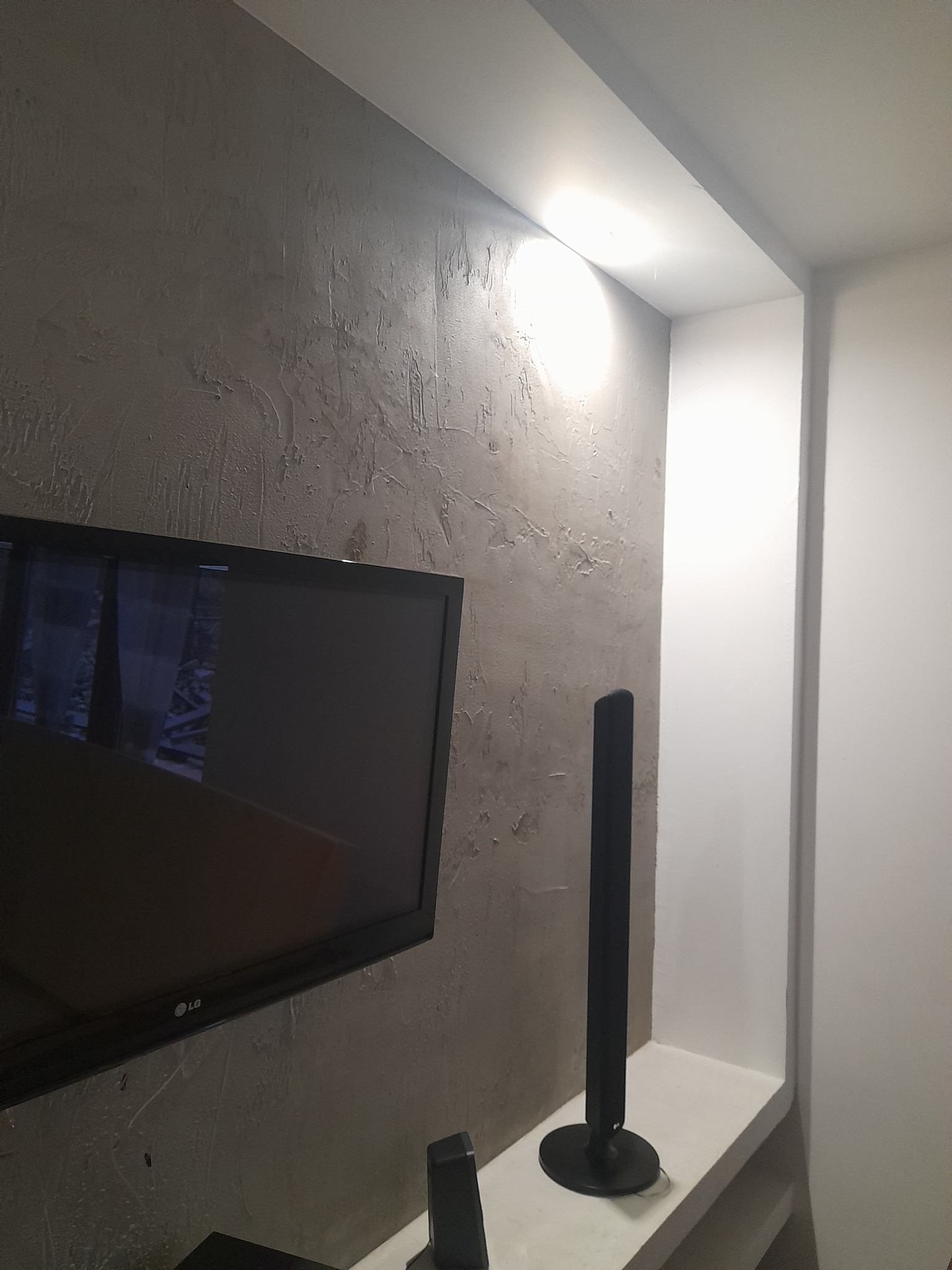 Ściana w salonie z dekoracyjną szarą strukturą, podświetlona górnym światłem, z widocznym telewizorem LG i głośnikiem.