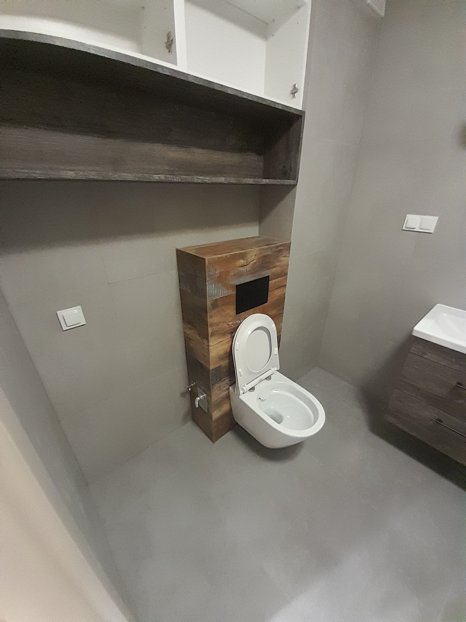 Nowoczesna łazienka z podwieszaną toaletą zabudowaną w drewnopodobnej obudowie, szara podłoga i ściany, wiszące szafki i szafka pod umywalką w podobnym stylu.