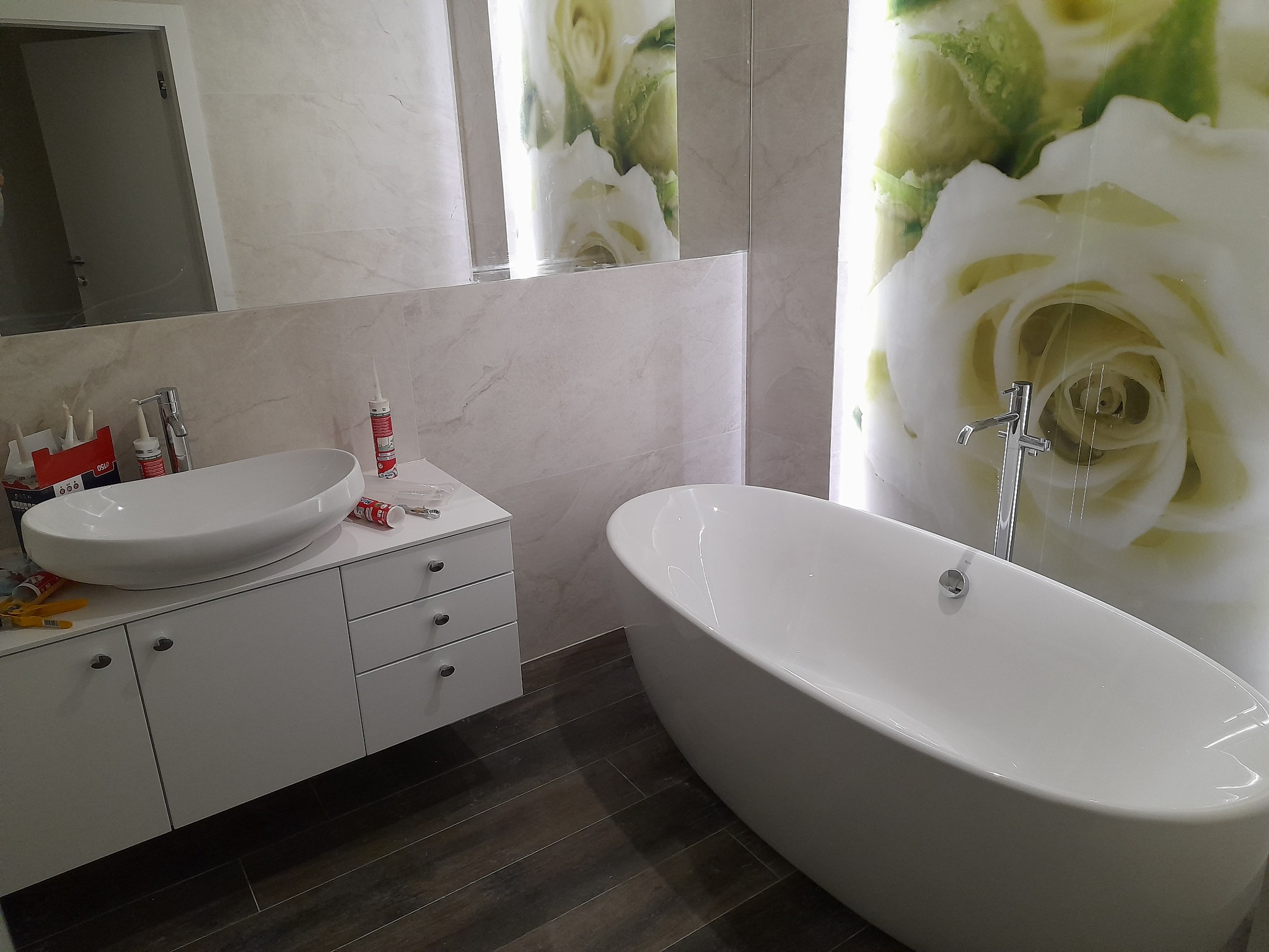 Nowoczesna łazienka z wolnostojącą wanną, umywalką nablatową na szafce z szufladami i panelami ściennymi z motywem białych róż. Widoczne tuby silikonu sanitarnego i narzędzia na blacie.