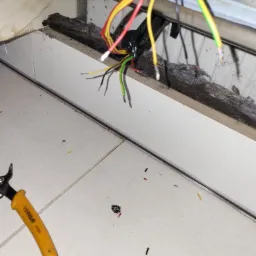 Podłączenie przewodów elektrycznych wewnątrz uszkodzonego sprzętu AGD, widoczne obcięte końcówki kabli, narzędzie do cięcia przewodów oraz zabrudzona podłoga z fragmentami izolacji.