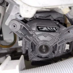 Szczegółowe ujęcie silnika elektrycznego wewnątrz pralki, widoczne elementy mocujące, przewody oraz fragment węża odpływowego.