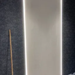 Ściana z gładzią w kolorze écru, oświetlona pionowymi pasami ledowymi, sąsiadująca z ciemną, teksturowaną tapetą. W rogu widoczny drewniany kij malarski.