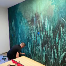 Mężczyzna w czarnej koszulce aplikuje tapetę z motywem trawy na ścianę, widoczne narzędzia na stole roboczym, niebieskie znaczniki na tapecie.