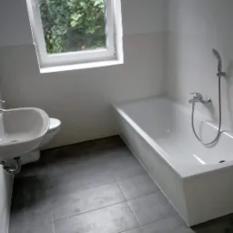 Jasna łazienka z szarymi płytkami podłogowymi, białą wanną i umywalką, okno z widokiem na zieleń.