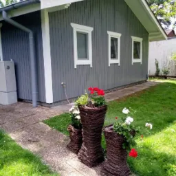 Szara elewacja domu z trzema oknami w białych ramach, obok ścieżki stoją trzy wiklinowe donice z czerwonymi i białymi kwiatami.