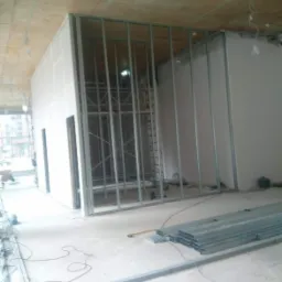 Montaż metalowej konstrukcji ścian działowych z płyt gipsowo-kartonowych w surowym wnętrzu, widoczne profile, płyty, narzędzia i przewody elektryczne na podłodze.