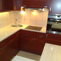 Kompaktowa kuchnia z ciemnymi, drewnianymi szafkami, jasnym blatem z imitacją kamienia, okrągłym zlewem i płytą indukcyjną; widoczny fragment piekarnika w zabudowie.