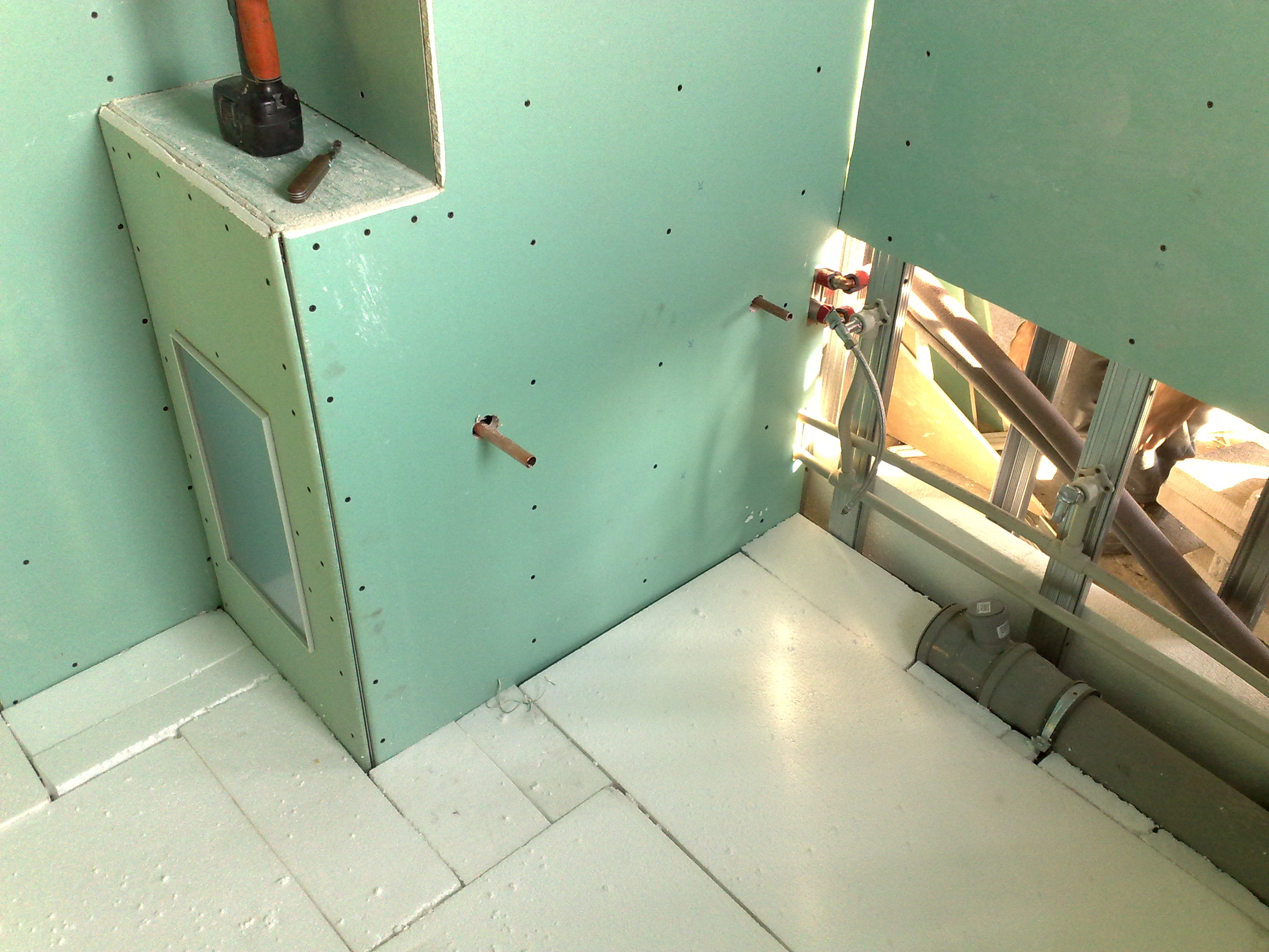 Poddasze w trakcie adaptacji: zielone płyty karton-gips na ścianach, izolacja podłogi z polistyrenu ekstrudowanego, widoczne rury instalacji wodnej i kanalizacyjnej, wiertarka i nóż na zabudowie.