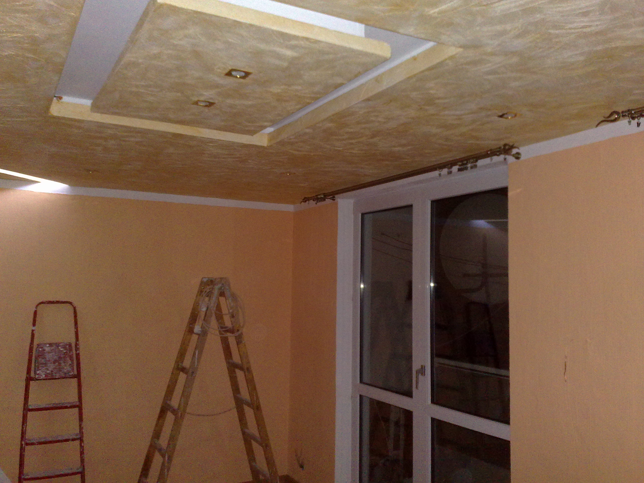 Wykończony sufit z płyty karton gips z wbudowanym oświetleniem w pokoju o pomarańczowych ścianach, widoczne dwie drabiny i okno balkonowe.