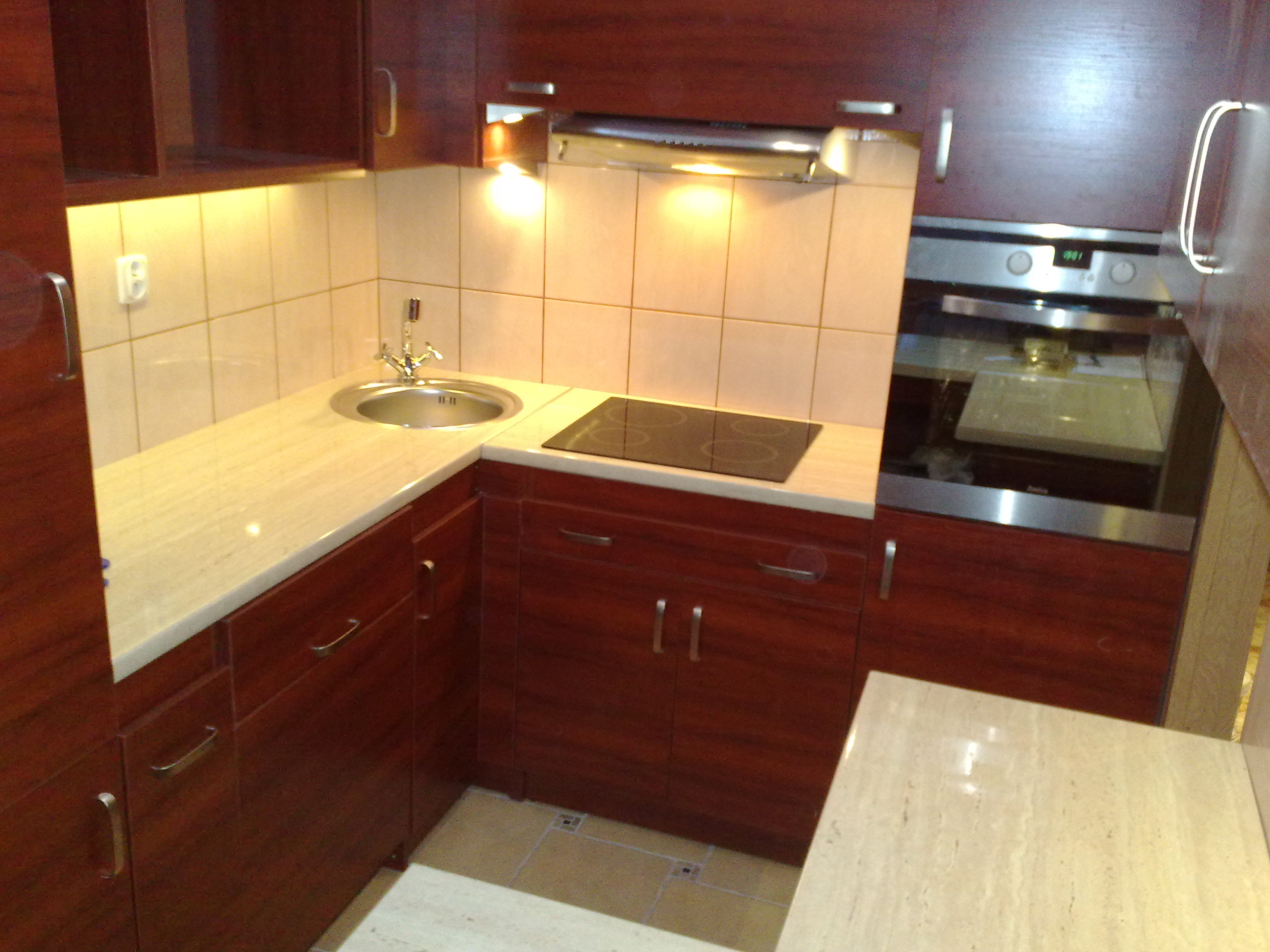 Kompaktowa kuchnia z ciemnymi, drewnianymi szafkami, jasnym blatem z imitacją kamienia, okrągłym zlewem i płytą indukcyjną; widoczny fragment piekarnika w zabudowie.