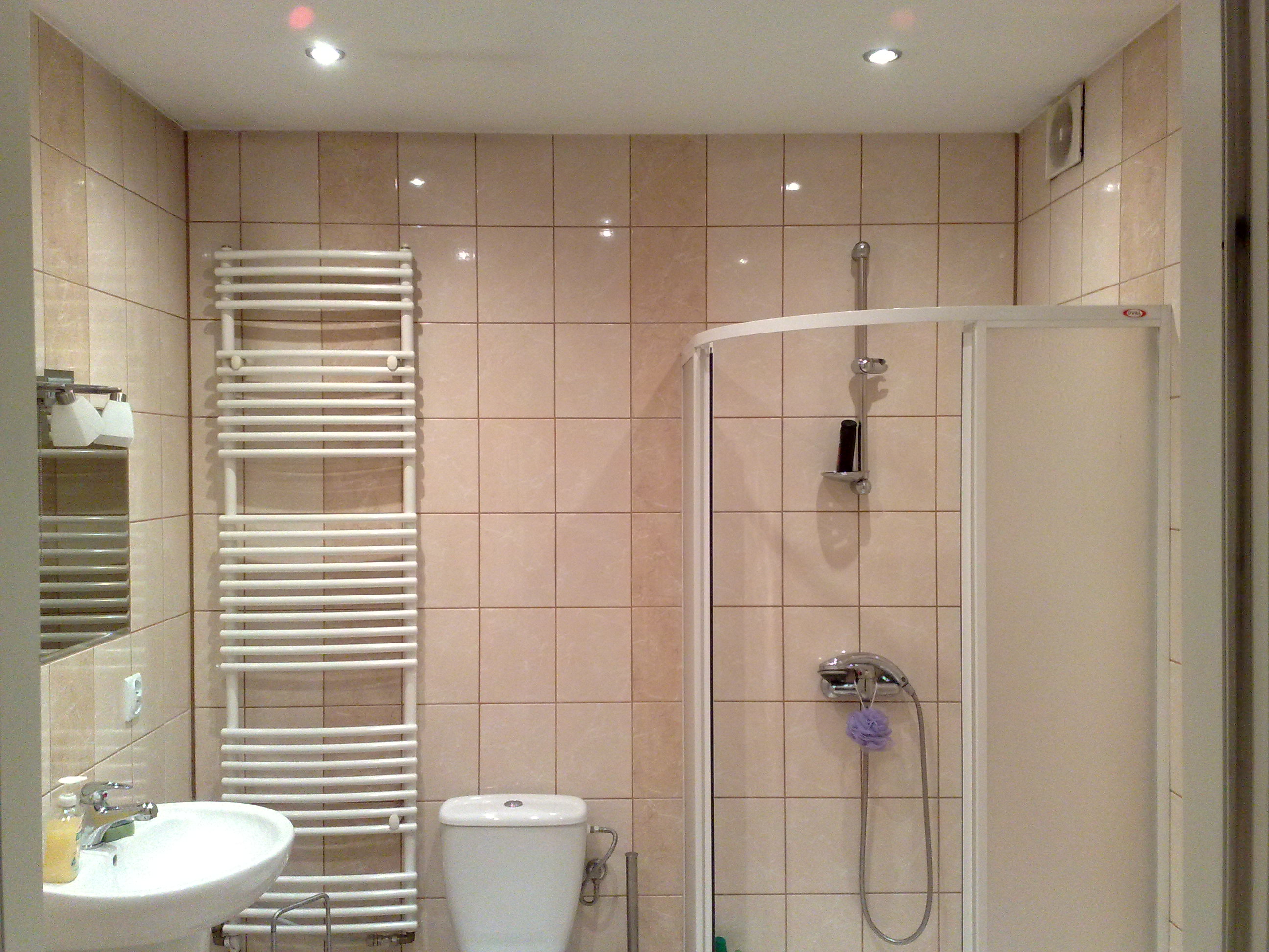 Jasna łazienka z beżowymi płytkami, wyposażona w białą umywalkę na postumencie, grzejnik drabinkowy, toaletę i kabinę prysznicową z półokrągłymi drzwiami.