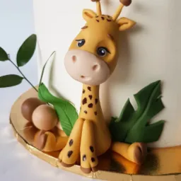 Tort zdobiony ręcznie wykonaną figurką żyrafy z lukru plastycznego, otoczonej liśćmi i brązowymi kulkami na złotej podstawie.