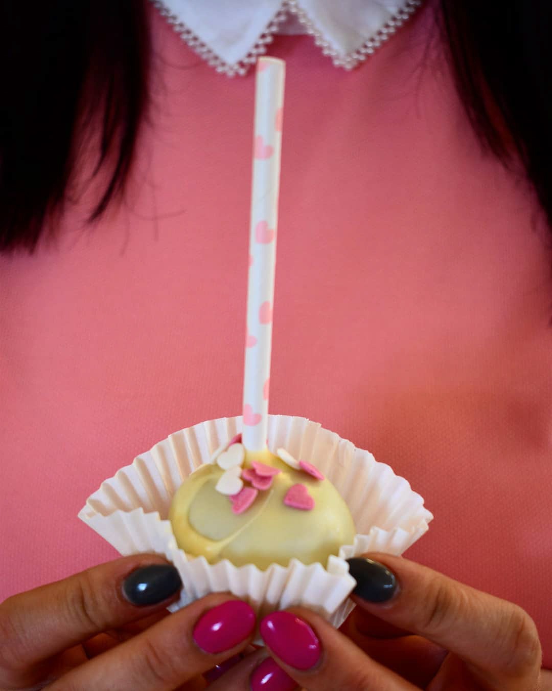 Pojedynczy cake pop w białej papierowej foremce, udekorowany różowymi i białymi serduszkami, trzymany przez osobę z różowo-czarnym manicurem na tle różowej bluzki z koronkowym kołnierzykiem.