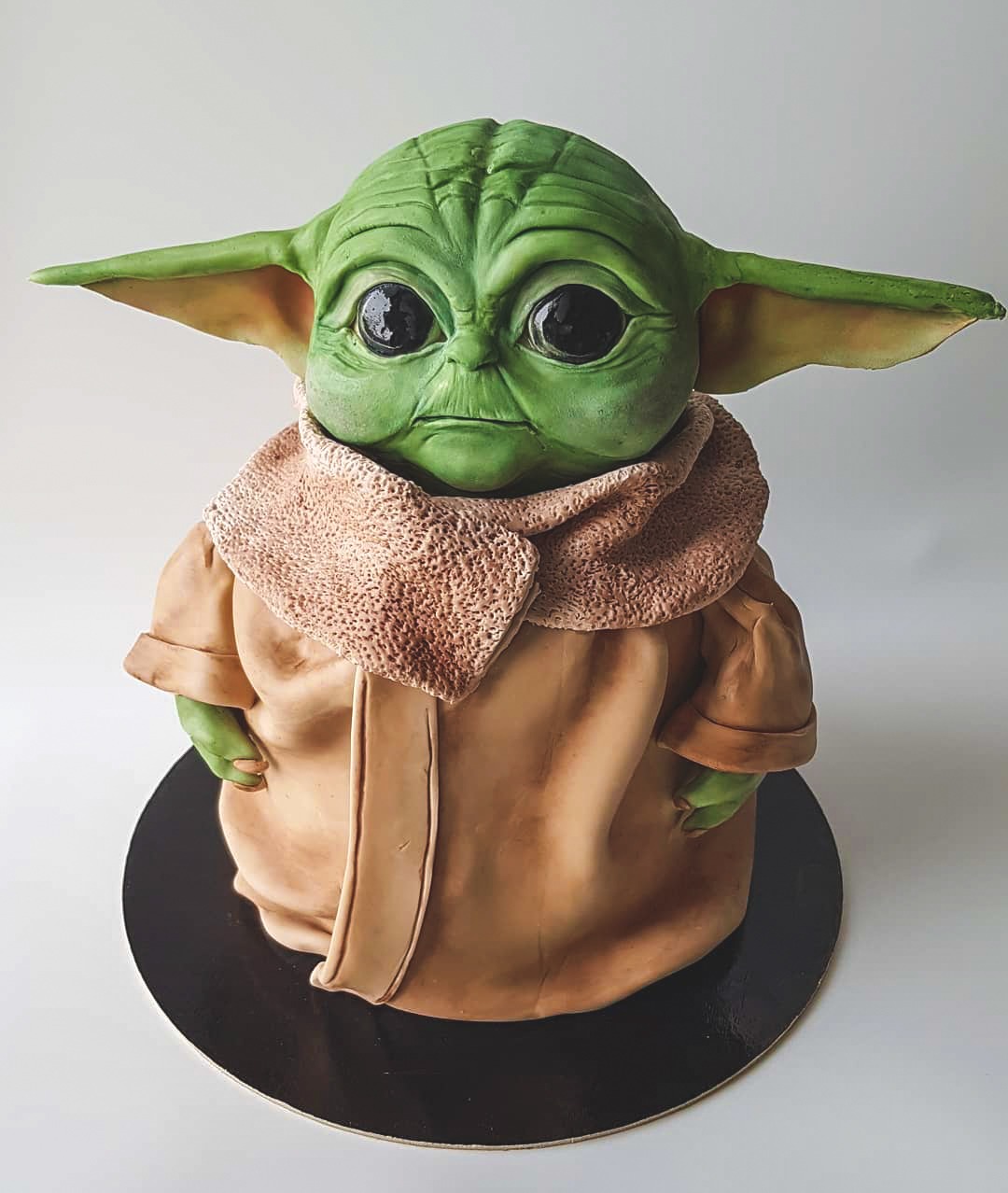Tort w kształcie postaci z Gwiezdnych Wojen, Baby Yoda, z zieloną skórą, dużymi czarnymi oczami i brązowym płaszczem, umieszczony na czarnym talerzu.