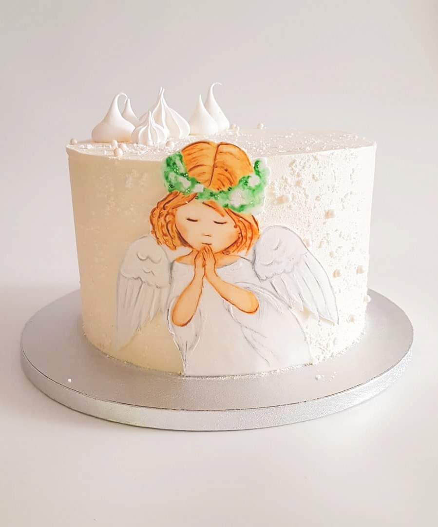 Tort z kremowym lukrem, dekorowany ręcznie malowanym aniołem z zielonym wiankiem i białymi skrzydłami, zwieńczony małymi bezami.