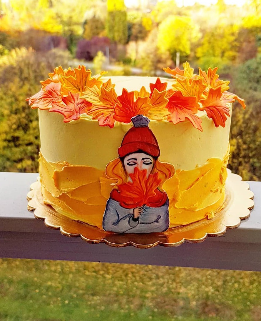 Tort z żółtym kremem, dekorowany pomarańczowymi liśćmi klonu i naklejką z wizerunkiem kobiety w czerwonej czapce, trzymającej liść, na złotym podkładzie, z rozmytym jesiennym tłem.