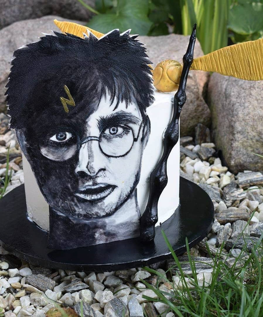 Okrągły tort z ręcznie malowanym portretem Harry'ego Pottera, złotym zniczem i czarną różdżką na tle kamieni i trawy.