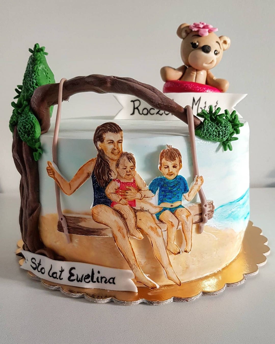 Tort urodzinowy z ręcznie malowaną sceną rodziny na huśtawce i misiem na szczycie, dekorowany zielonymi elementami przypominającymi rośliny.