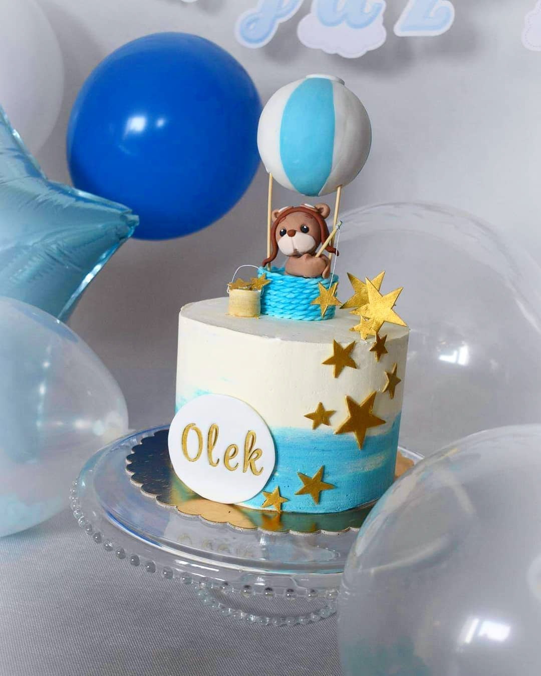 Tort urodzinowy dla chłopca z motywem misia w balonie, dekorowany złotymi gwiazdkami i napisem 'Olek', otoczony niebieskimi i białymi balonami.