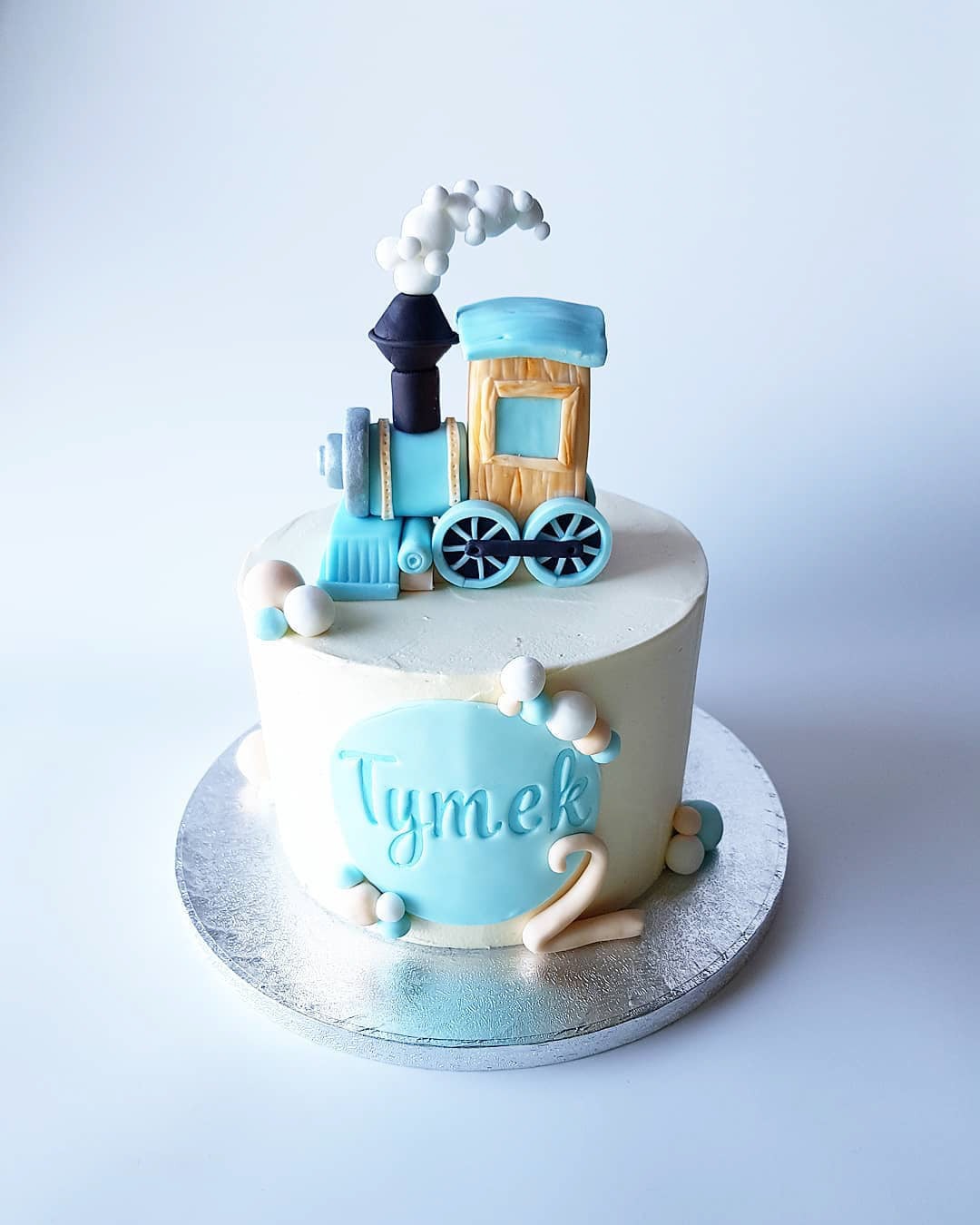 Tort urodzinowy dla dziecka z dekoracją w kształcie błękitnego pociągu i napisem 'Tymek 2' na srebrnym podkładzie.
