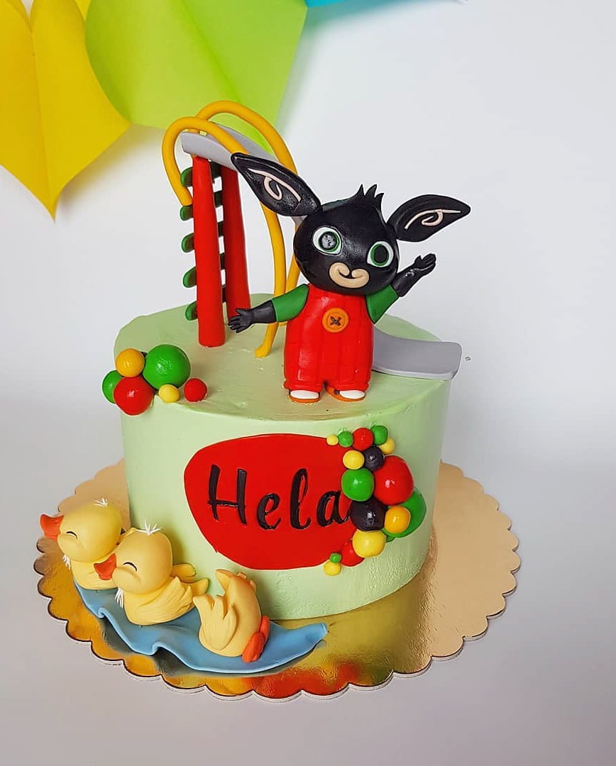Tort urodzinowy z motywem Binga, dekorowany figurkami kaczuszek i napisem 'Hela', umieszczony na złotej podkładce.
