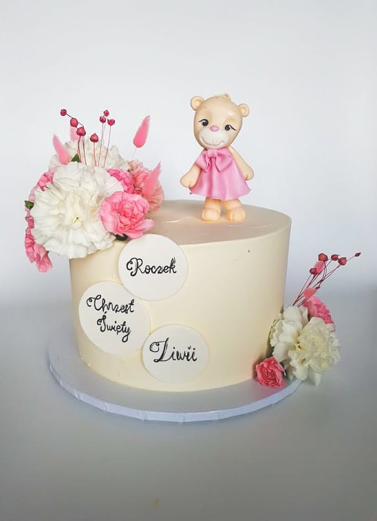 Tort okolicznościowy z kremowym lukrem, dekorowany kwiatami w odcieniach bieli i różu, figurką misia w różowej sukience oraz trzema okrągłymi etykietami z napisami: Roczek, Chrzest Święty, Liwii.