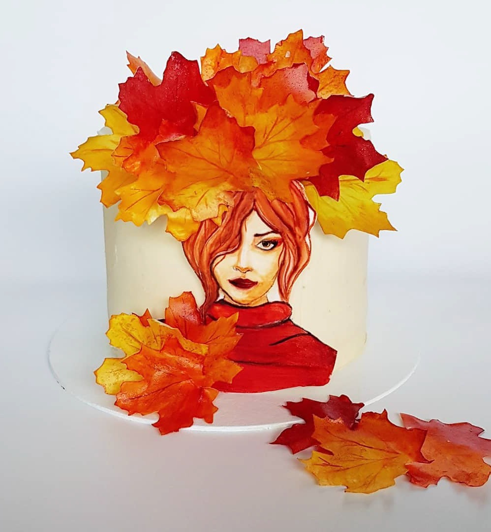 Tort zdobiony ręcznie malowanym portretem kobiety w czerwonym swetrze, otoczony jadalnymi liśćmi klonu w odcieniach czerwieni, pomarańczy i żółci, imitującymi jesienną aurę.