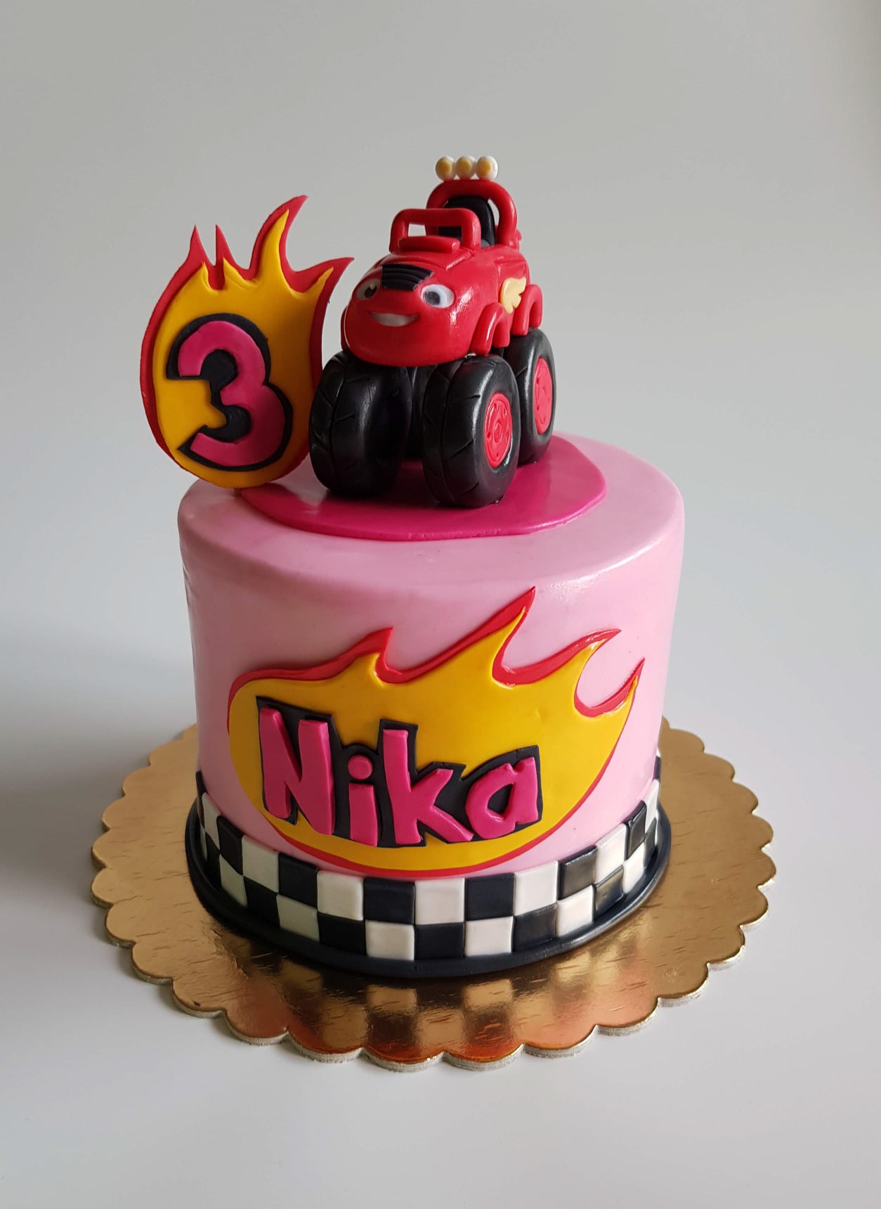 Różowy tort urodzinowy z motywem monster trucka i imieniem Nika, numer trzy w płomieniach, szachownicowa obwódka.
