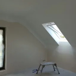 Poddasze w trakcie prac wykończeniowych: świeżo położone gładzie na skośnych ścianach, okno dachowe wpuszczające jasne światło, drabinka malarska na środku pomieszczenia, okno zabezpieczone folią.