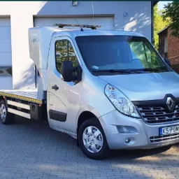 Srebrny samochód Renault Master z platformą lawety, zaparkowany na kostce brukowej przed budynkiem z białymi drzwiami garażowymi, tablica rejestracyjna z napisem 'K5 POMOC'.