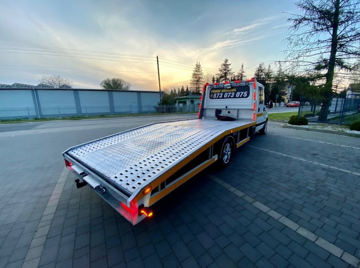 Pusta laweta pomocy drogowej z numerem telefonu na zabudowie, zaparkowana na parkingu z kostki brukowej, w tle budynek i drzewa pod wieczornym niebem.