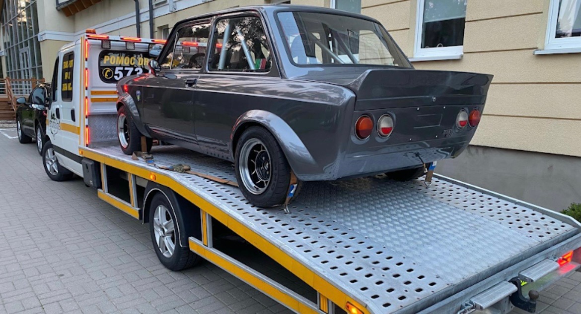 Szary, tuningowany Fiat 128 z klatką bezpieczeństwa przewożony na platformie lawety. Auto zabezpieczone pasami transportowymi.