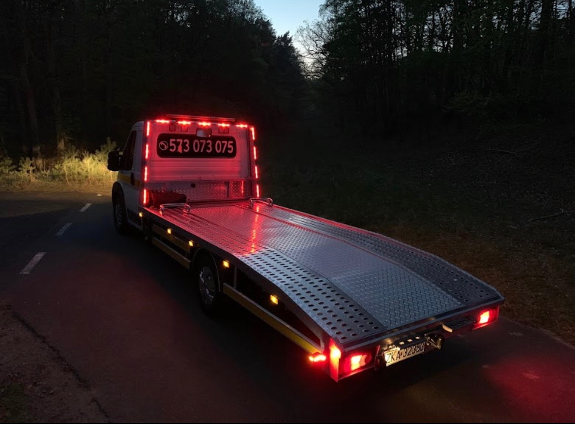 Laweta z podświetleniem LED i numerem telefonu na zabudowie, stojąca na drodze w lesie o zmierzchu.