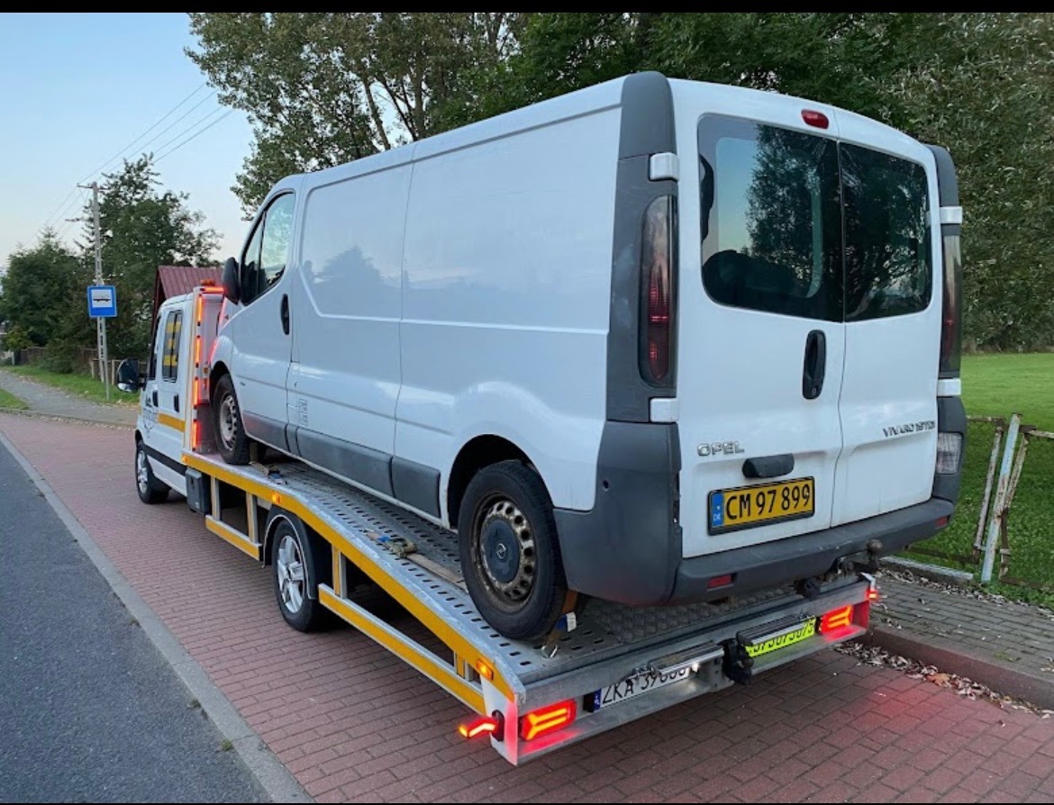 Biały bus Opel Vivaro na żółtej lawecie na tle zieleni i drogi. Widoczny numer rejestracyjny busa.