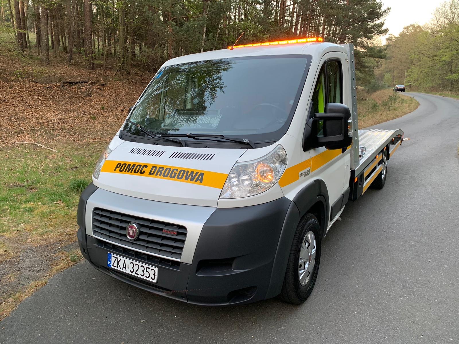 Biała laweta Fiat z napisem 'Pomoc Drogowa' i włączonymi pomarańczowymi światłami ostrzegawczymi stoi na asfaltowej drodze w lesie.