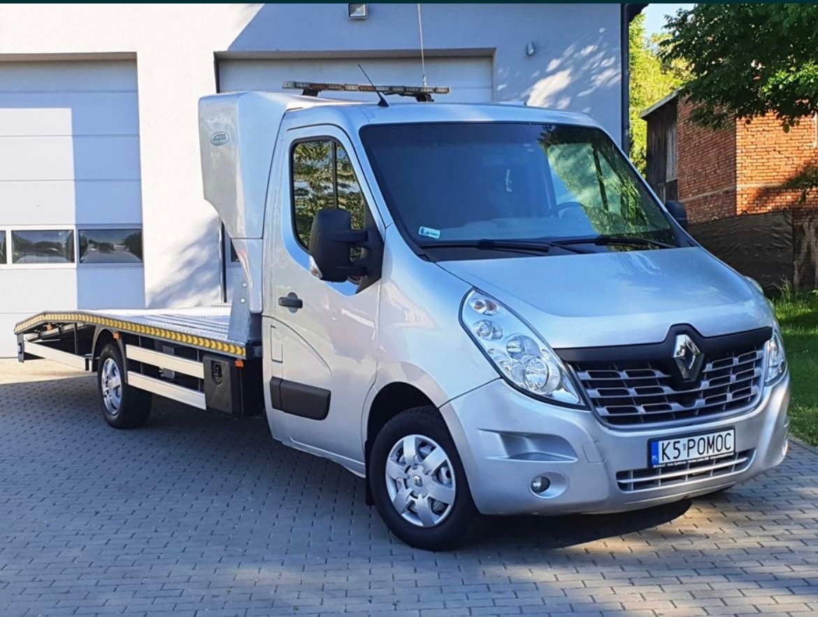 Srebrny samochód Renault Master z platformą lawety, zaparkowany na kostce brukowej przed budynkiem z białymi drzwiami garażowymi, tablica rejestracyjna z napisem 'K5 POMOC'.
