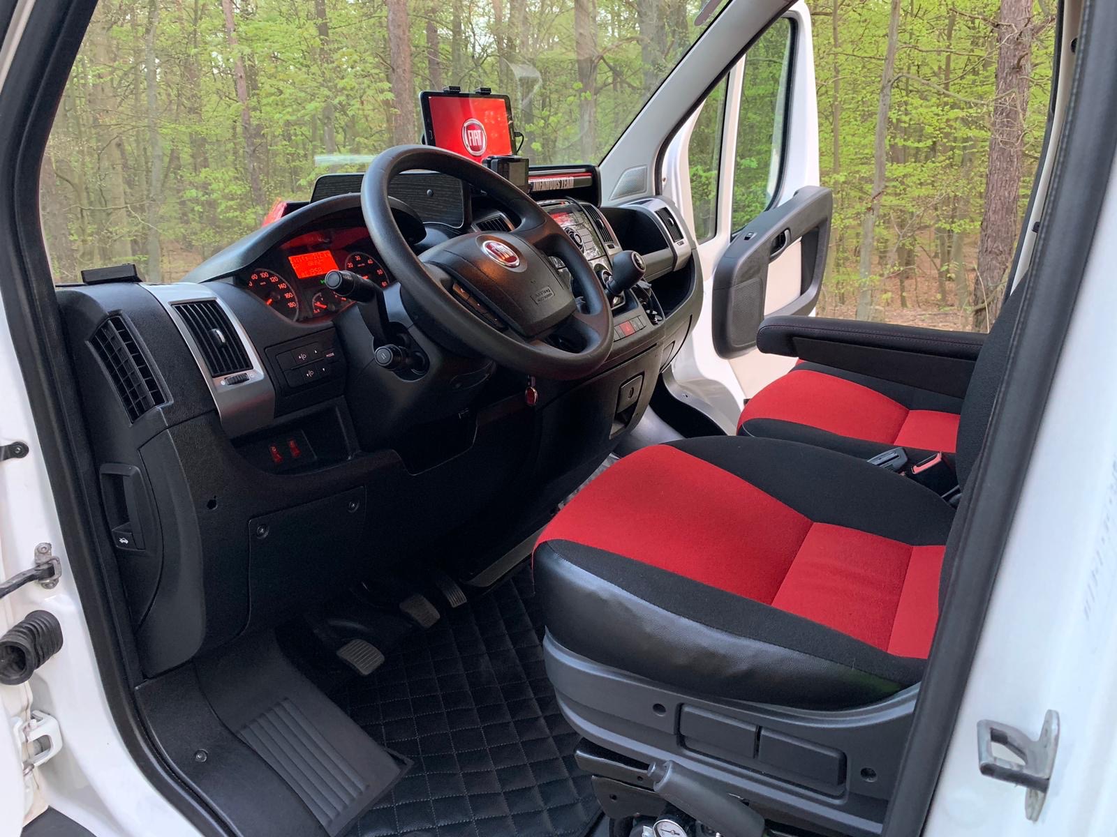 Wnętrze białego Fiata Ducato z czerwono-czarnymi fotelami, widok z otwartych drzwi na deskę rozdzielczą, kierownicę i tablet z logo Fiata, w tle zielony las.
