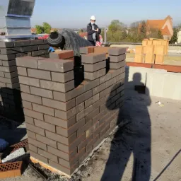 Wrocław: Murarz w trakcie wznoszenia komina z cegły klinkierowej na dachu budynku, widoczny fragment miasta w tle.