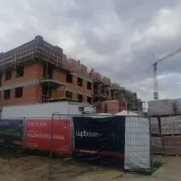 Wznoszenie wielopiętrowego budynku z cegły, widoczne rusztowania, palety z materiałami budowlanymi i żuraw budowlany na tle pochmurnego nieba, ogrodzenie z banerami reklamowymi