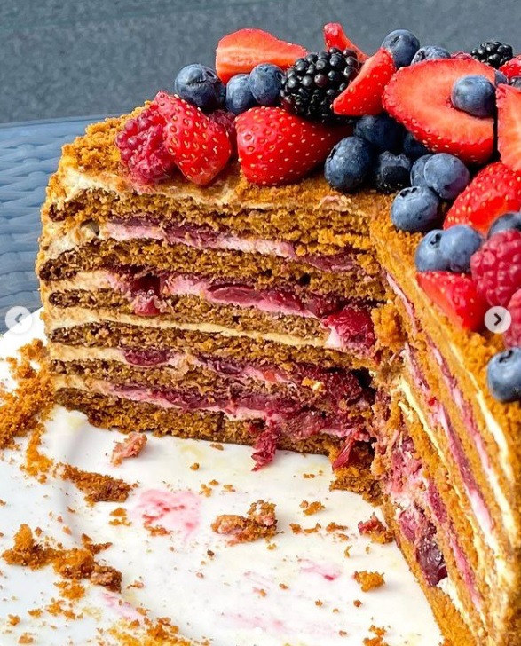 Przekrój tortu miodowego z warstwami wiśni i dekoracją z truskawek, malin, borówek i jeżyn, widoczne okruchy na talerzu.