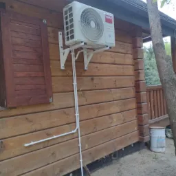 Zewnętrzna jednostka klimatyzacji marki Vivax zamontowana na drewnianej ścianie domu z brązowymi okiennicami, widoczne białe rurki odprowadzające skropliny.