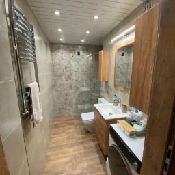 Wyremontowana łazienka z drewnianymi akcentami: szafka pod umywalką, lustro w drewnianej ramie, grzejnik drabinkowy, płytki imitujące kamień na ścianach, drewnopodobne panele na podłodze, pralka...