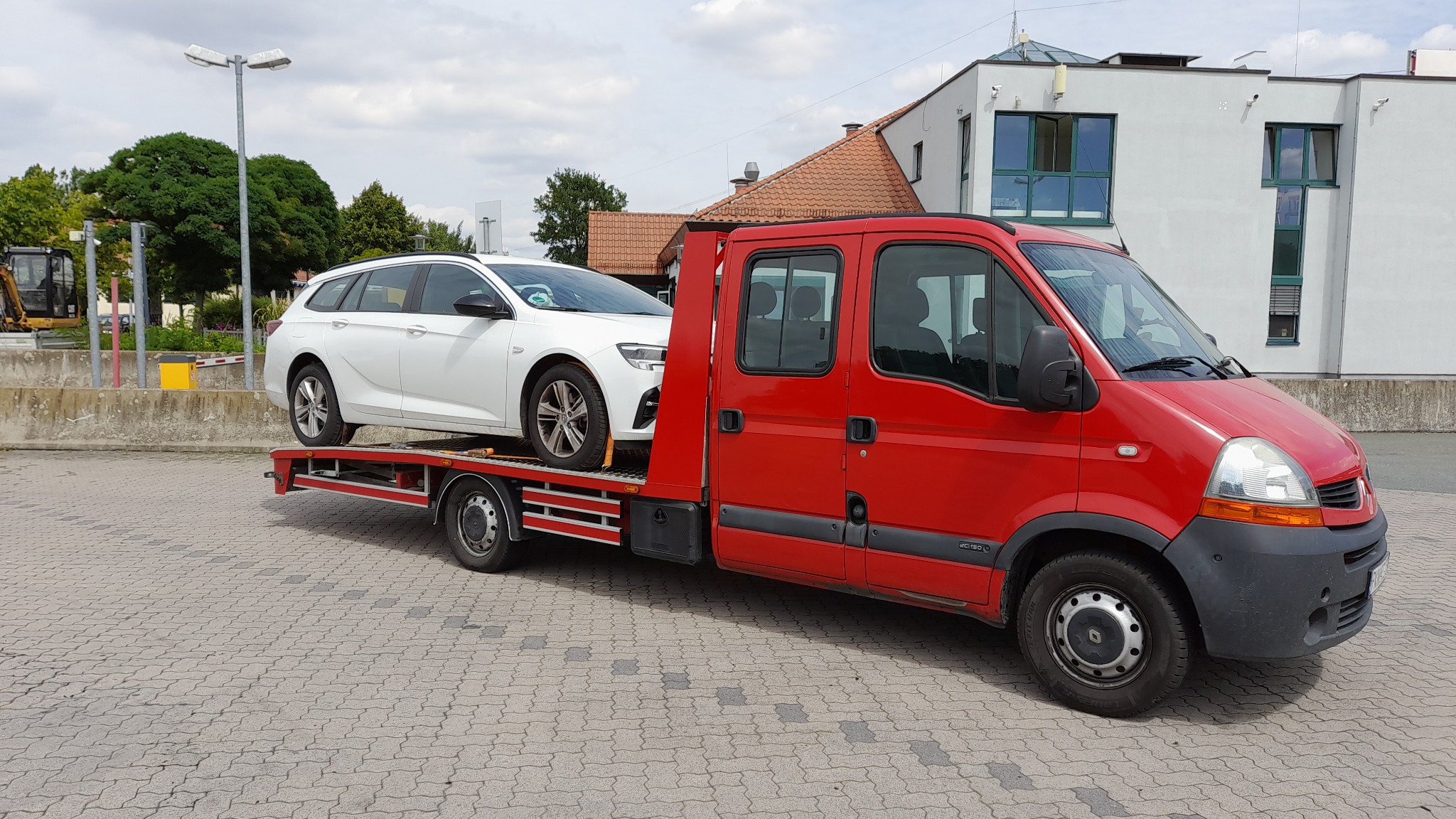 Czerwona laweta z białym samochodem osobowym typu kombi na platformie, zaparkowana na brukowanym placu, w tle budynek i zieleń.