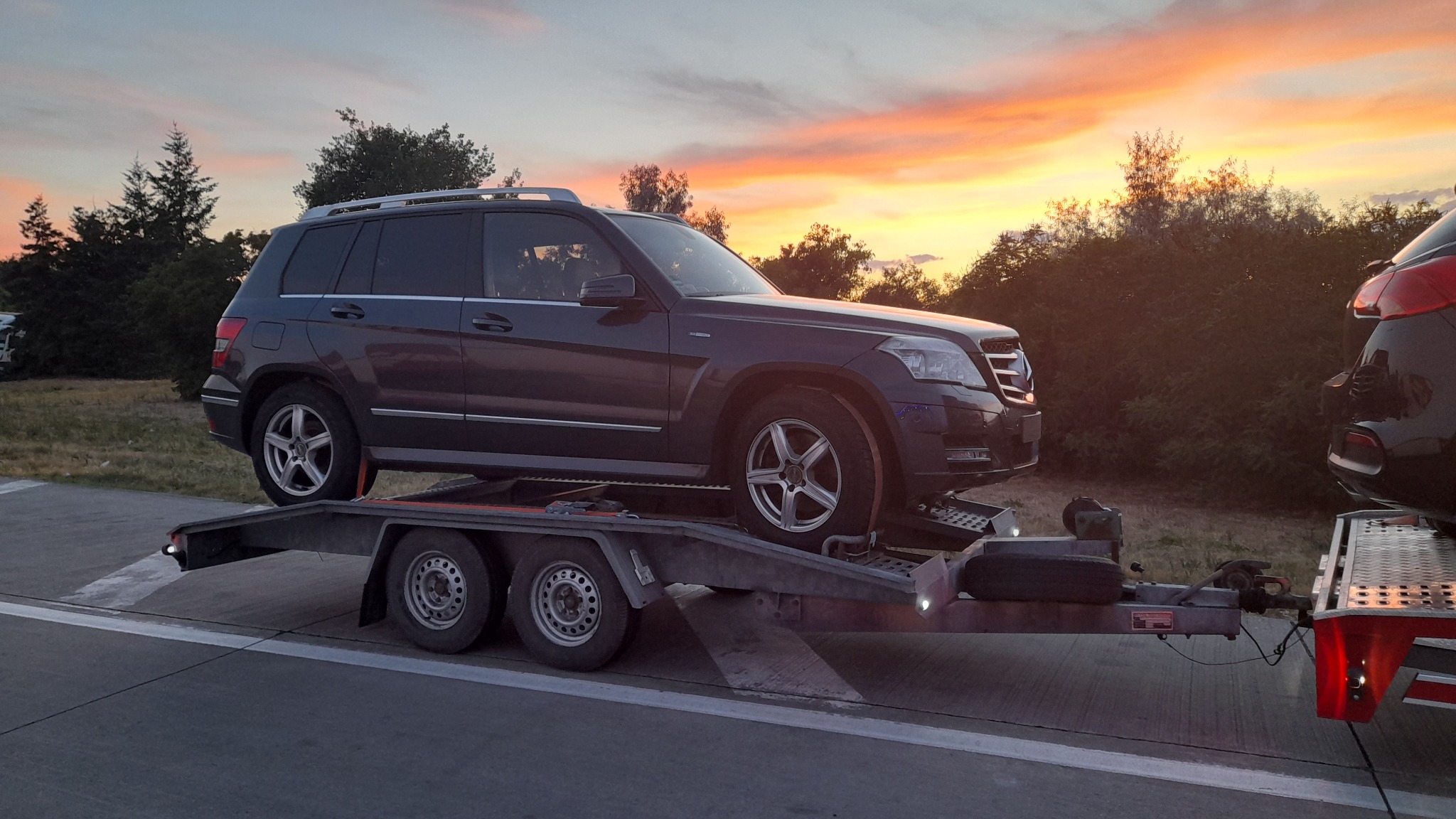 Ciemnoszary samochód SUV na lawecie z dwoma osiami, z zachodem słońca w tle. Widoczny drugi pojazd na lawecie z tyłu.