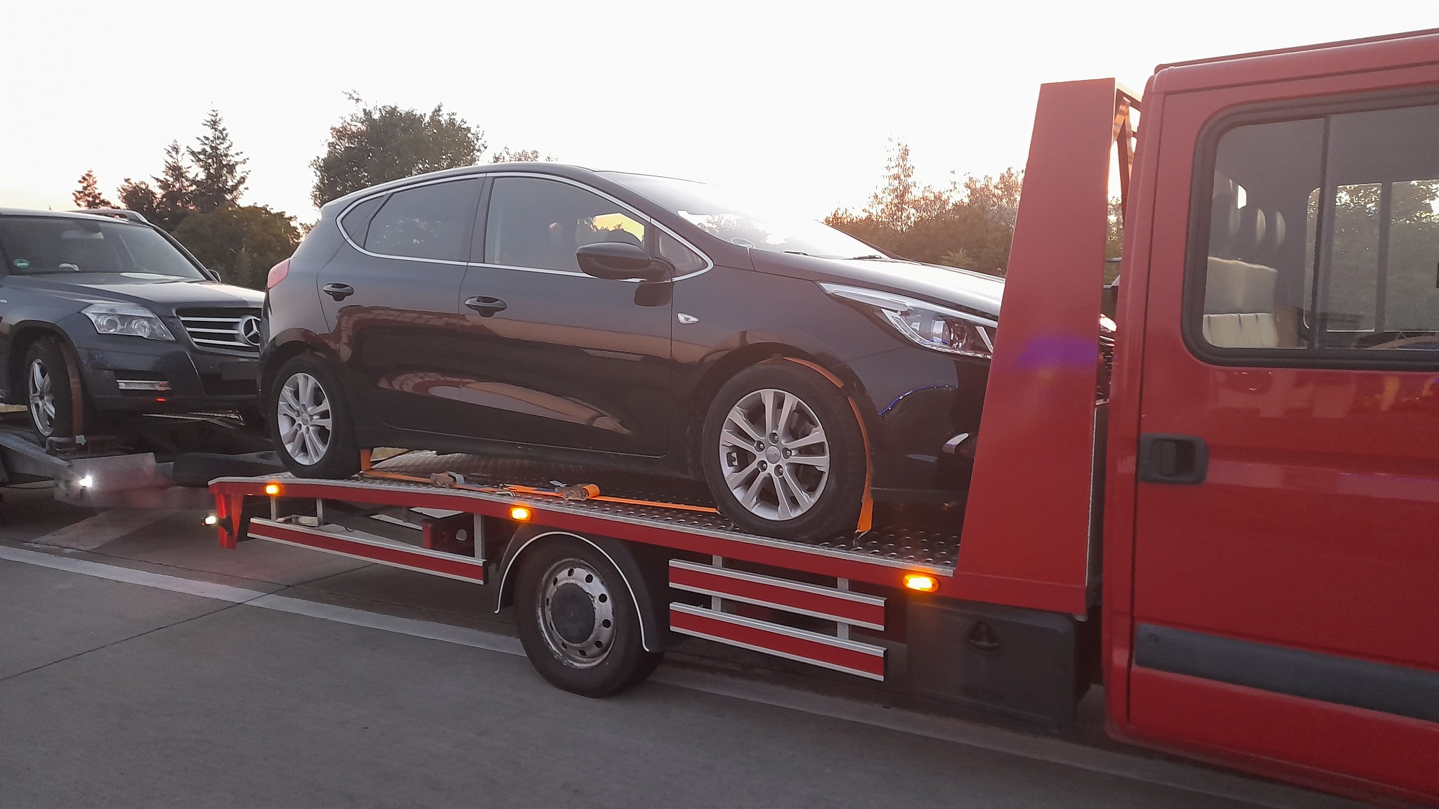 Dwa samochody, Kia Ceed i Mercedes GLK, przewożone na platformie lawety w trakcie zachodu słońca.