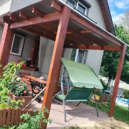 Zadaszona drewniana pergola z metalowym dachem, pod nią stół z ławkami, huśtawka ogrodowa z zielonym daszkiem, obok rabaty kwiatowe i drewniane płotki.