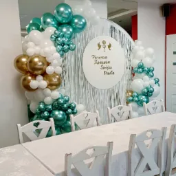 Dekoracja sali na Pierwszą Komunię Świętą z białymi krzesłami z wyciętymi sercami, stołem nakrytym białym obrusem w kwiaty oraz balonami białymi, złotymi i turkusowymi, z białym okrągłym banerem...